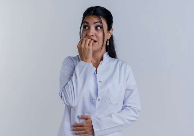 Ansioso joven doctora vistiendo bata médica mirando de lado poniendo la mano en el vientre y mordiendo los dedos aislados