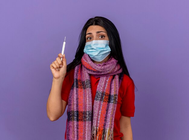 Ansioso joven enferma caucásica vistiendo máscara y bufanda sosteniendo termómetro aislado en la pared púrpura con espacio de copia