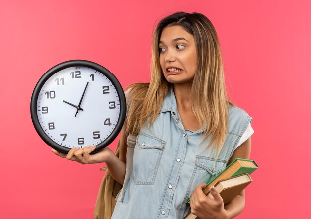 Ansioso joven estudiante bonita vestida con bolsa trasera con reloj y libros mirando el reloj aislado en rosa