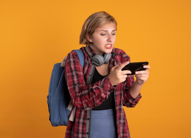 Ansioso joven estudiante eslavo con auriculares con mochila sostiene y mira el teléfono