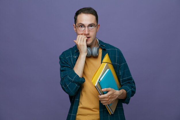 Ansioso joven estudiante masculino con anteojos auriculares alrededor del cuello sosteniendo una carpeta de bloc de notas mirando a la cámara mordiendo los dedos aislado en un fondo morado