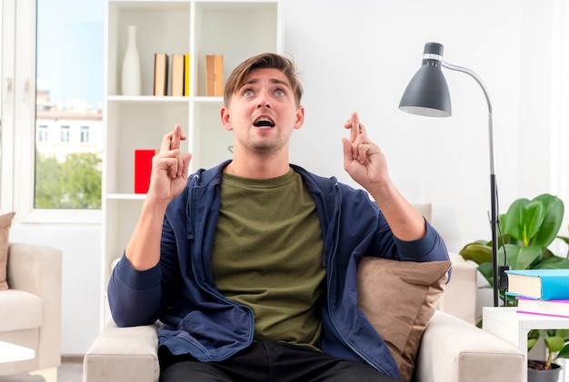 Ansioso joven guapo rubio se sienta en un sillón cruzando los dedos mirando hacia arriba