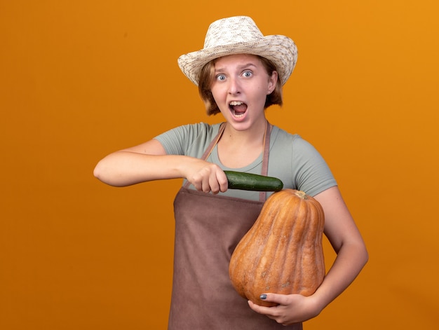 Ansioso joven jardinero eslavo vistiendo sombrero de jardinería sosteniendo pepino y calabaza en naranja