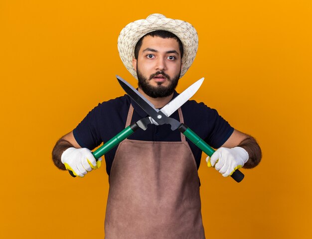 Ansioso joven jardinero varón caucásico vistiendo gorro de jardinería y guantes sosteniendo tijeras de jardinería aislado en la pared naranja con espacio de copia