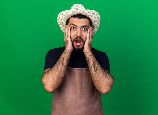 Ansioso joven jardinero varón caucásico vistiendo sombrero de jardinería pone las manos en el rostro aislado en la pared verde con espacio de copia