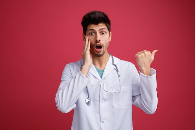 Ansioso joven médico masculino con uniforme médico y estetoscopio alrededor del cuello mirando a la cámara apuntando hacia un lado manteniendo la mano en la cara aislada en el fondo rojo