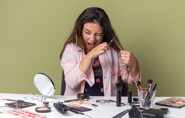 Ansioso joven morena sentada a la mesa con herramientas de maquillaje cortándose el pelo con unas tijeras