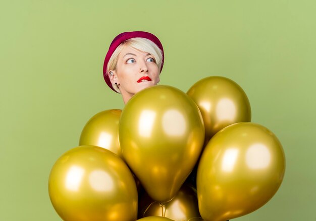 Ansioso joven rubia fiestera con gorro de fiesta de pie detrás de globos mirando el labio mordedor lateral aislado sobre fondo verde oliva