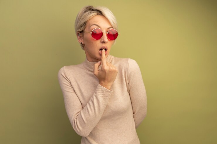 Foto gratuito ansioso joven rubia con gafas de sol manteniendo la mano detrás de la espalda poniendo el dedo en el labio