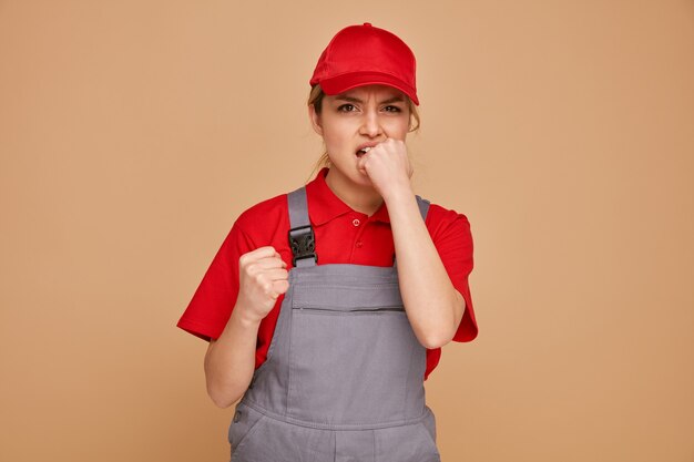 Ansioso joven trabajador de la construcción con gorra y uniforme apretando el puño mordiendo otro puño