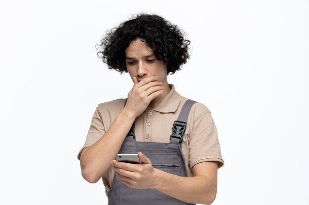 Ansioso joven trabajador de la construcción vistiendo uniforme manteniendo la mano en la boca sosteniendo y mirando el teléfono móvil aislado sobre fondo blanco.