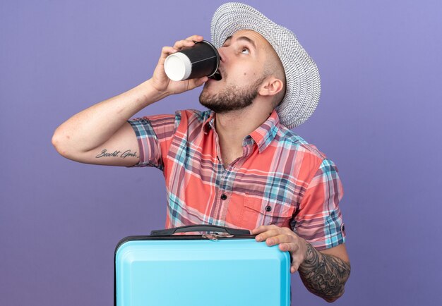 Ansioso joven viajero caucásico hombre con sombrero de playa de paja bebiendo de taza de papel de pie detrás de la maleta aislado sobre fondo púrpura con espacio de copia