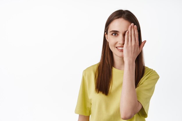 Antes después efecto belleza. Una joven sonriente cubre la mitad de la cara con la mano, se ve feliz con un ojo, parada en una camiseta contra el fondo blanco