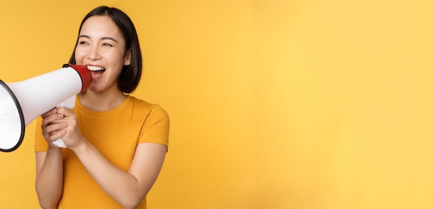 Anuncio Mujer asiática feliz gritando fuerte en megáfono reclutando protestando con altavoz en manos de pie sobre fondo amarillo