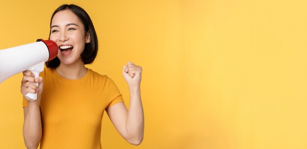 Anuncio Mujer asiática feliz gritando fuerte en megáfono reclutando protestando con altavoz en manos de pie sobre fondo amarillo