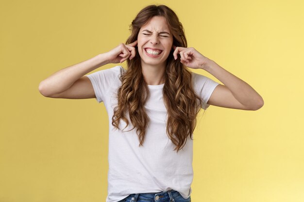 Apaga esta horrible música. Muchacha que hace la cara irónica cerró las orejas los dedos índices tapados los orificios para los oídos cerrar los ojos apretar los dientes disgustado molesto ruidoso terrible ruido irritado de vecinos fondo amarillo.