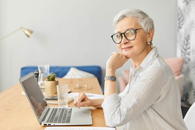 Aparatos electrónicos modernos, concepto de ocupación, edad y madurez. Vista lateral de la atractiva mujer autónoma de mediana edad con estilo en gafas sentado frente a la computadora portátil abierta, trabajando en la oficina en casa