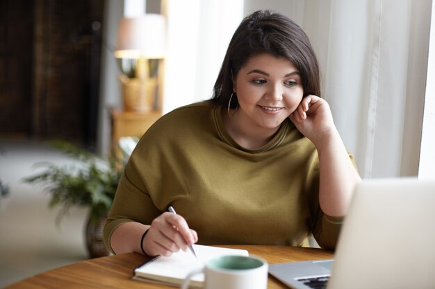 Aparatos electrónicos modernos, concepto de trabajo y ocupación. Mujer morena joven elegante linda con exceso de peso usando la computadora portátil para el trabajo remoto, mirando la pantalla, sosteniendo la pluma, escribiendo