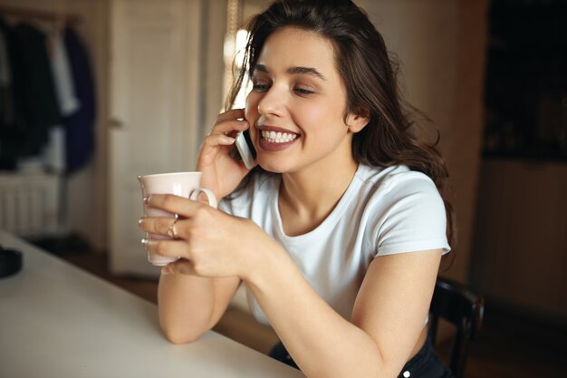 Aparatos electrónicos, personas, concepto de comodidad y relajación. Chica guapa con linda sonrisa disfrutando de una llamada telefónica, hablando con su novio mientras bebe café por la mañana, sentada en la mesa blanca con taza