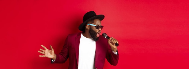 Foto gratuita apasionado cantante masculino negro actuando contra un fondo rojo cantando en una fiesta con micrófono