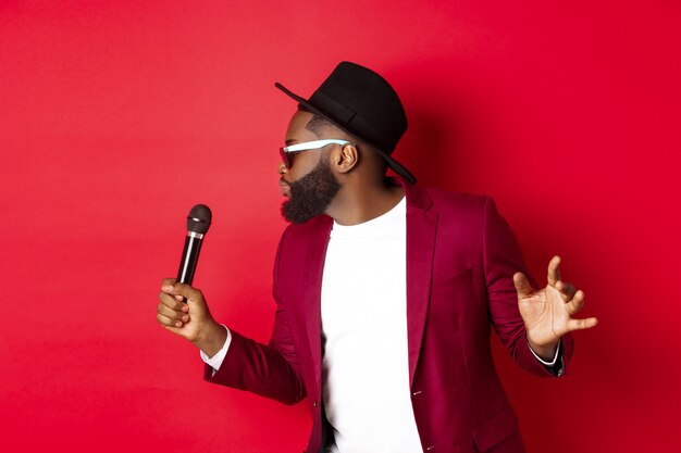 Apasionado cantante masculino negro actuando sobre fondo rojo, cantando en el micrófono, vistiendo traje de fiesta, de pie sobre fondo rojo.