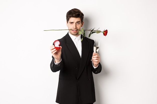 Apasionado joven en traje haciendo una propuesta, sosteniendo una rosa en los dientes y una copa de champán, mostrando el anillo de compromiso, pidiendo casarse con él, de pie contra el fondo blanco.