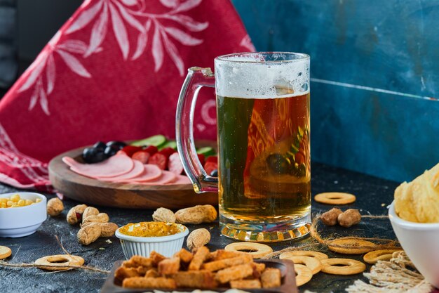 Aperitivos variados, patatas fritas, un plato de salchichas y un vaso de cerveza sobre una superficie oscura.