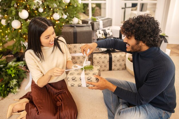 Apertura de regalos. Linda mujer asiática joven de pelo largo y hombre indio sonriente abriendo regalo de navidad juntos tocando la cinta en la caja sentado en el piso cerca del árbol de navidad