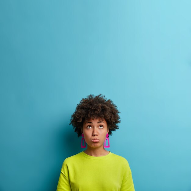 Apesadumbrada mujer de piel oscura se ve con expresión de tristeza frustrada arriba, infeliz por los problemas, usa camiseta verde y aretes, pared azul con espacio en blanco vacío