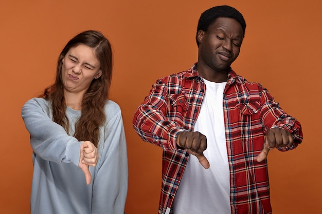 Esto apesta. Retrato de una joven pareja interracial emocional, varón africano y mujer caucásica mostrando los pulgares hacia abajo gesto, disgustado con la mala comida o el hedor, suprimiendo los vómitos. Disgusto y disgusto