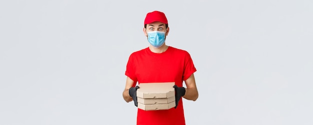 Aplicación de entrega de alimentos compras en línea sin contacto y concepto covid19 Mensajero sorprendido con máscara facial uniforme roja y guantes se ven impresionados traer a los clientes cajas de pizza