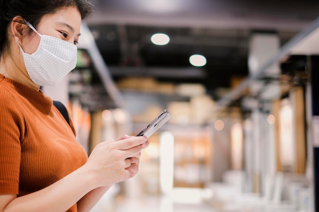 Aplicación de teléfono inteligente que busca una mujer asiática que usa una máscara facial que escribe a mano una comunicación tect de teléfono inteligente en un centro comercial de grandes almacenes