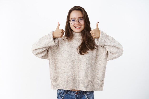 Apoyo y optimismo alegre agradable morena europea con gafas y suéter mostrando los pulgares hacia arriba impresionado y encantado, que le gusta el producto dando una respuesta positiva, recomendándolo sobre la pared gris.