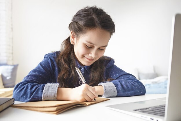 Aprendizaje, educación, ocio, afición y tecnologías modernas. Alegre adolescente positiva sentada en un escritorio en su habitación, tomando notas en su diario