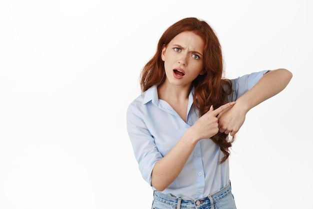 Apresúrate. Chica pelirroja molesta señalando el gesto de la muñeca del reloj, diciendo que se mueva más rápido, llegamos tarde, diciendo la hora, de pie en blusa contra fondo blanco.