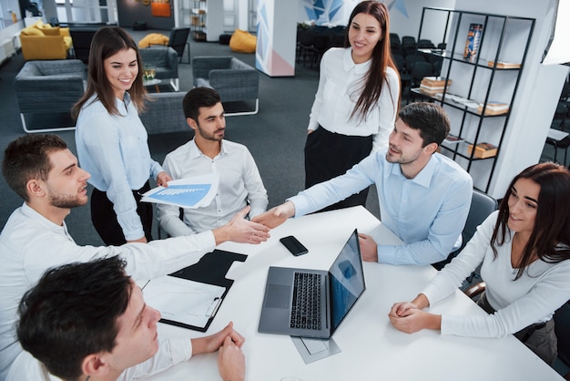 Apretón de manos para el acuerdo de éxito. Vista superior de los trabajadores de oficina en ropa clásica sentados cerca de la mesa usando una computadora portátil y documentos