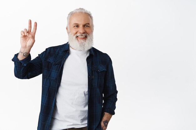 Un apuesto anciano hipster muestra el signo de la paz y sonríe feliz. Un viejo con barba y tatuajes hace un signo de apoyo de pie sobre un fondo blanco.