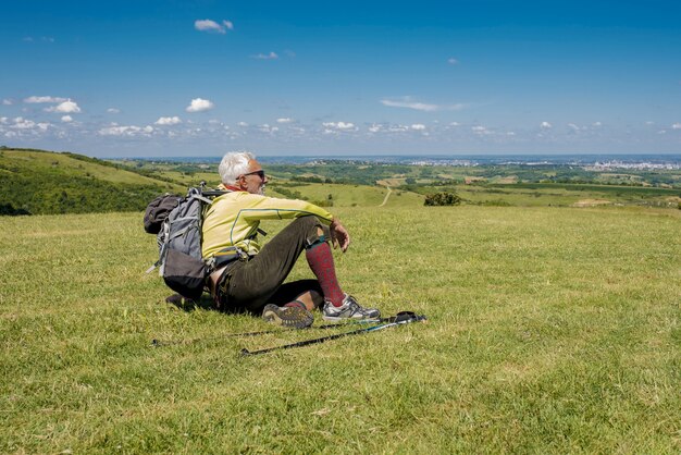 Apuesto anciano sentado en un prado y mirando relajante después de una caminata