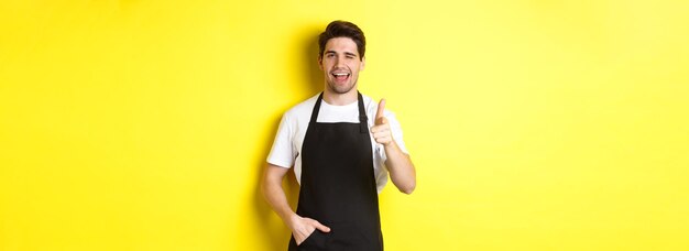 Apuesto barista en delantal negro da la bienvenida a los invitados a la cafetería apuntando con el dedo y guiñando el pie standi