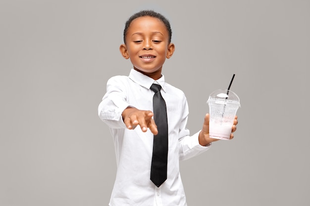 Apuesto colegial negro con camisa y corbata sosteniendo un vaso de plástico transparente bebiendo un batido de proteína energética saludable con expresión facial feliz y complacida. Salud y alimentación