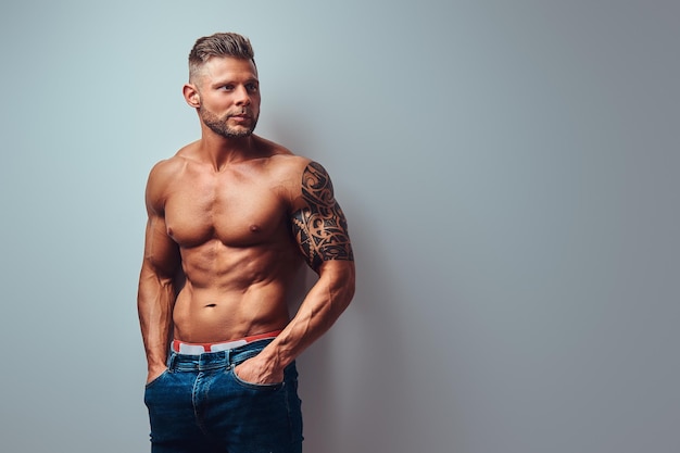 Un apuesto culturista sin camisa con un elegante corte de pelo y barba, con un tatuaje en el brazo, posando en un estudio. Aislado en un fondo gris.