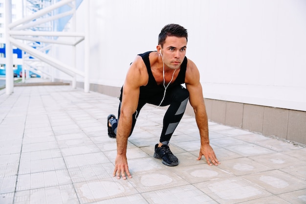 Apuesto deportista muscular en auriculares en posición de partida, preparándose para correr