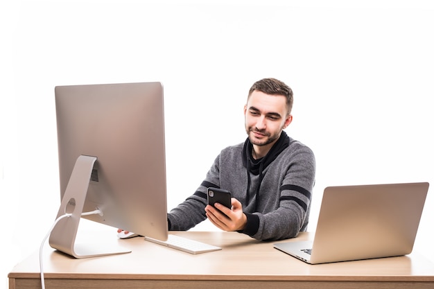 Apuesto empresario sentado detrás de su escritorio con computadora portátil y pc y escribiendo en el teléfono móvil aislado en blanco