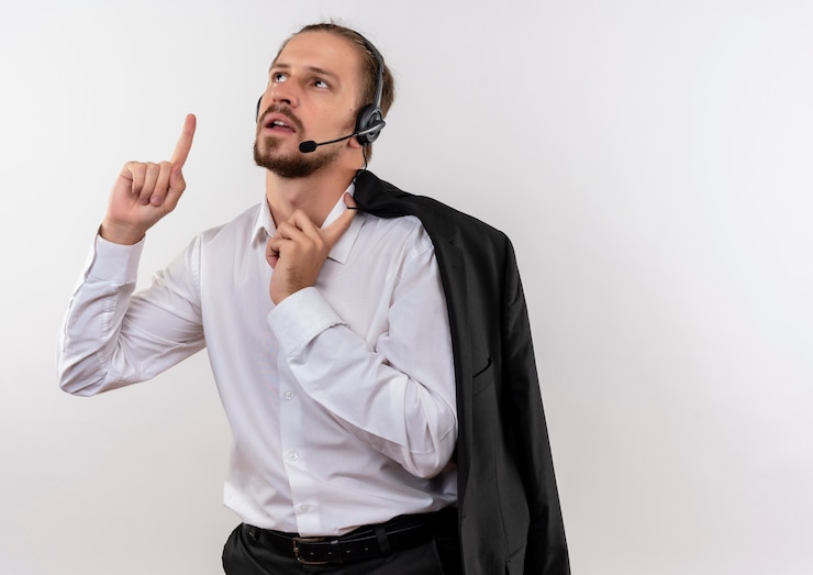 Foto gratuito apuesto empresario sosteniendo la chaqueta sobre el hombro con auriculares con un micrófono mirando hacia arriba con el dedo índice de pie sobre fondo blanco.