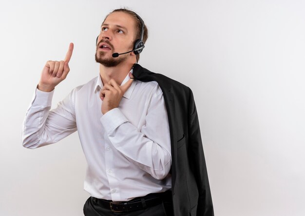 Apuesto empresario sosteniendo la chaqueta sobre el hombro con auriculares con un micrófono mirando hacia arriba con el dedo índice de pie sobre fondo blanco.