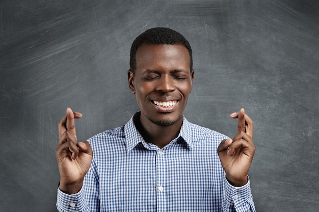 Apuesto estudiante africano en camisa a cuadros cruzando los dedos en ambas manos y manteniendo los ojos cerrados, pidiendo deseos, esperando lo mejor y orando por milagro, deseando aprobar exámenes con altas calificaciones