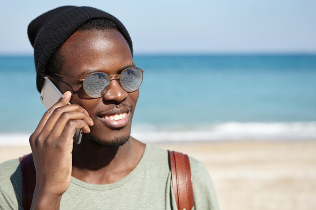 Apuesto estudiante afroamericano feliz con sombreros redondos y sombrero sonriendo ampliamente, hablando por teléfono móvil con sus padres, diciendo que lo está haciendo bien mientras viaja solo durante las vacaciones de verano