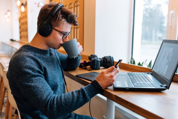 Apuesto fotógrafo bebiendo una taza de té en el café, mira el teléfono móvil