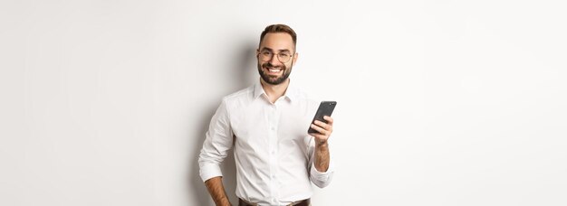 Apuesto gerente usando un teléfono inteligente y sonriendo complacido enviando un mensaje de texto de pie sobre el respaldo blanco