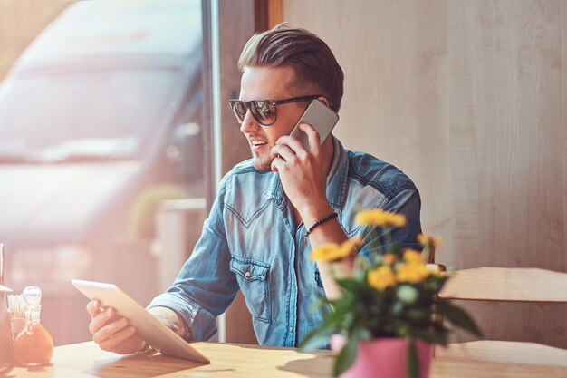 Un apuesto hipster con un elegante corte de pelo y barba se sienta en una mesa en un café al borde de la carretera, habla por teléfono y sostiene una tableta.